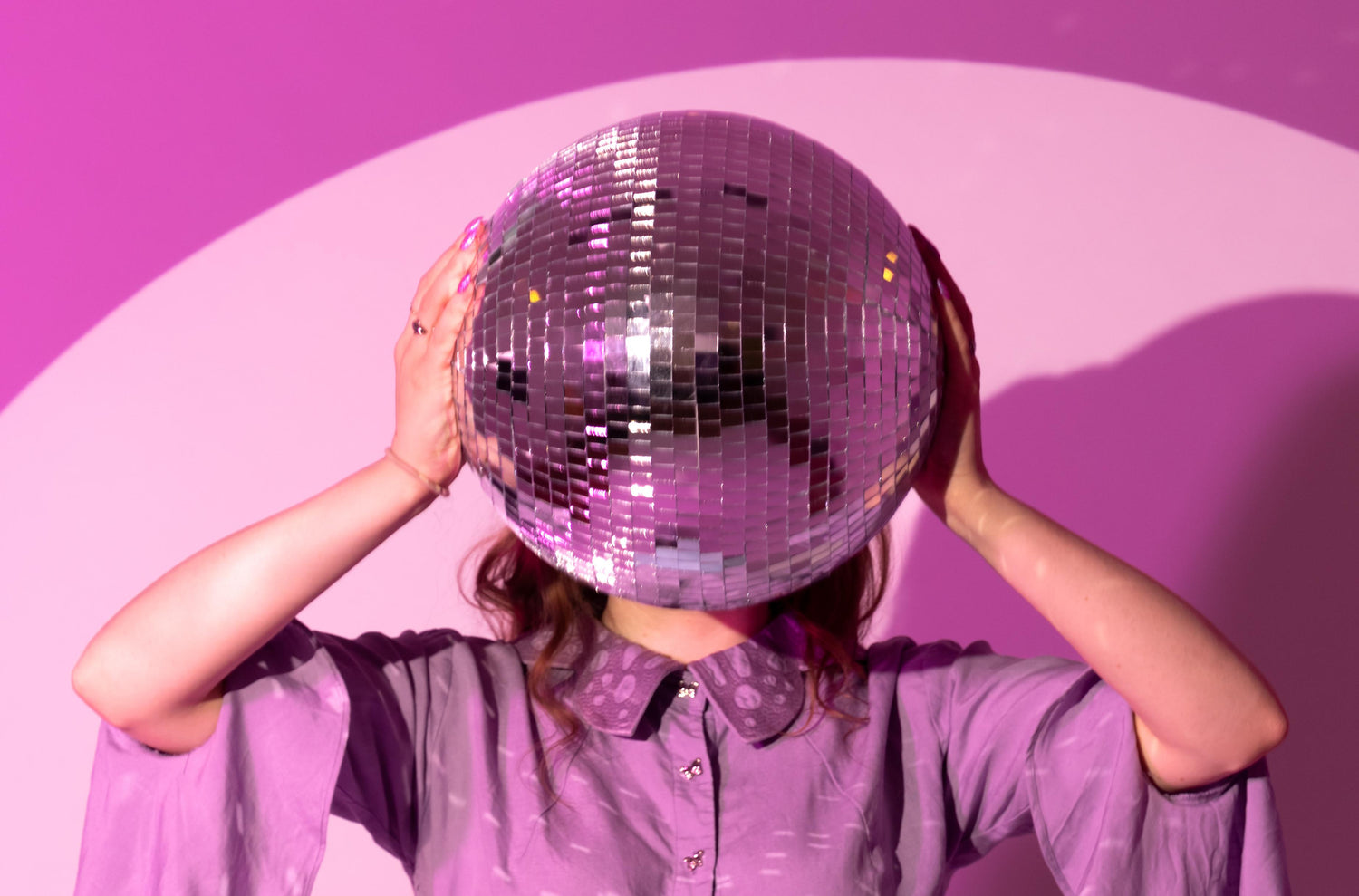 Person holding a disco ball in front of their face against a pink background